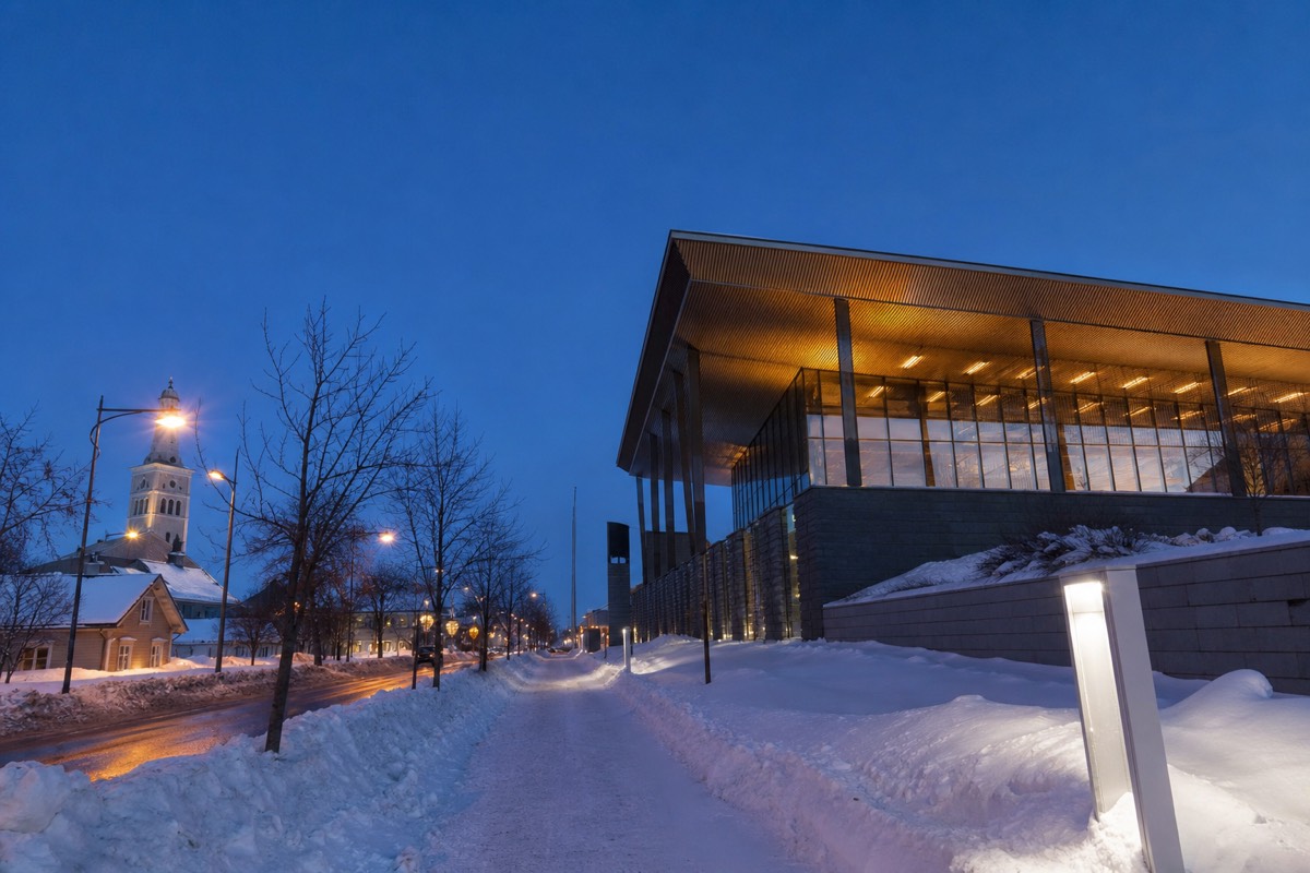 A snow-covered Finnish town street with a lit modern museum-style building at blue hour, snowbanks along the pavement