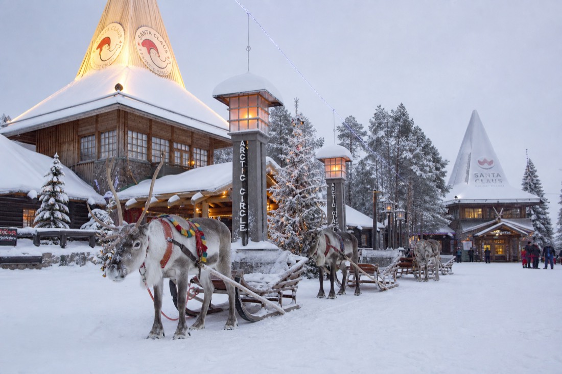 Santa Claus Village in Rovaniemi with reindeer outside wooden buildings in deep snow, warm lit windows, Arctic setting