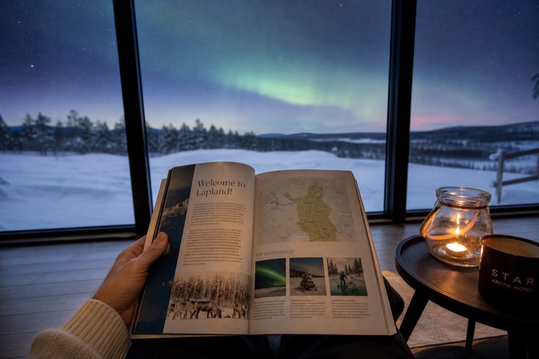 A person holding open a Lapland travel guidebook, sitting by a window overlooking snowy landscape and distant aurora