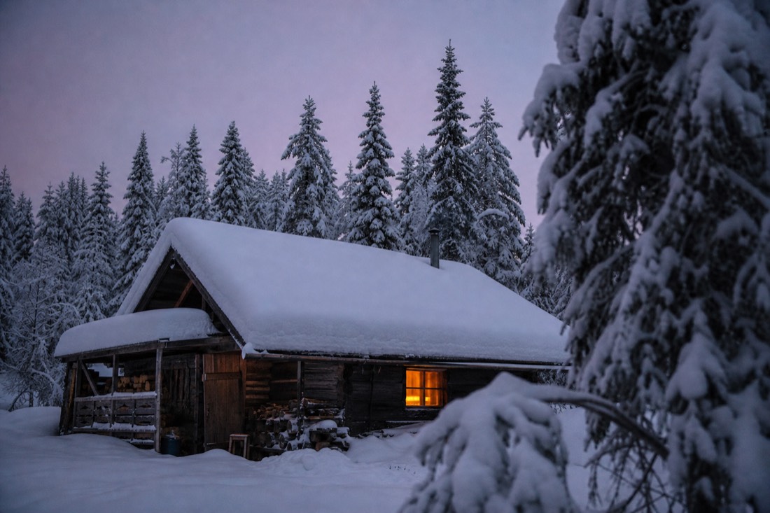 A wooden Lapland cabin under thick snow at dusk, one window lit warm