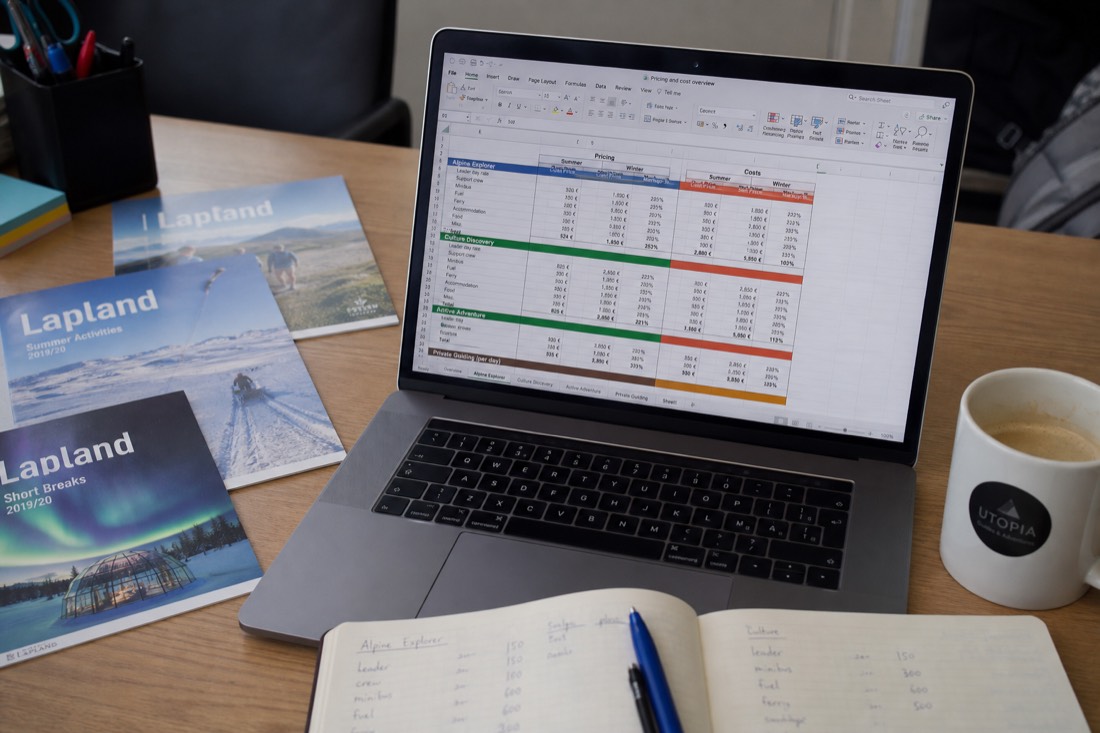 A spreadsheet with travel costs and pricing data on a desk with Lapland travel brochures and a coffee cup