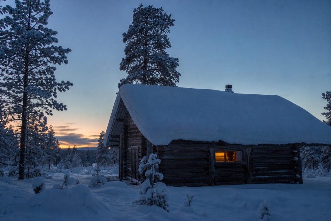 A wooden Lapland cabin under thick snow at dusk, one window lit warm