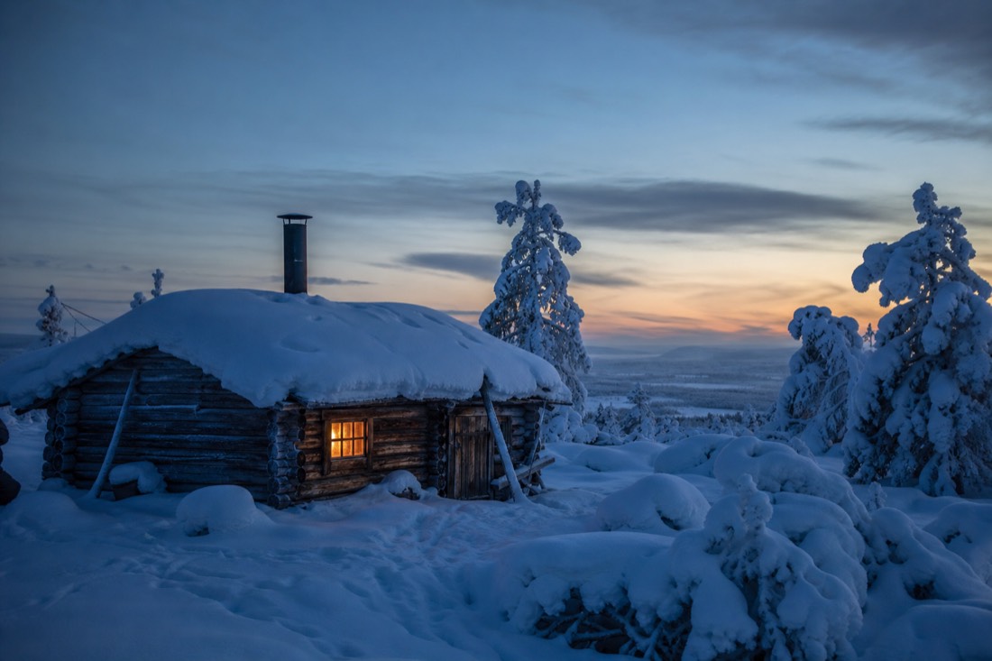 A wooden Lapland cabin under thick snow at dusk, one window lit warm